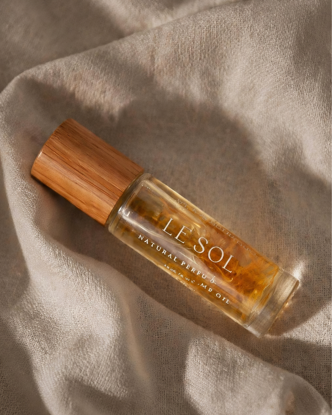 A clear glass perfume bottle with a light brown bamboo cap, containing amber-colored fragrance oil. The bottle is labeled 'LE SOL NATURAL PERFUME' and is placed on a draped beige linen fabric under warm, directional sunlight.