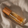A clear glass perfume bottle with a light brown bamboo cap, containing amber-colored fragrance oil. The bottle is labeled 'LE SOL NATURAL PERFUME' and is placed on a draped beige linen fabric under warm, directional sunlight.