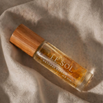 A clear glass perfume bottle with a light brown bamboo cap, containing amber-colored fragrance oil. The bottle is labeled 'LE SOL NATURAL PERFUME' and is placed on a draped beige linen fabric under warm, directional sunlight.